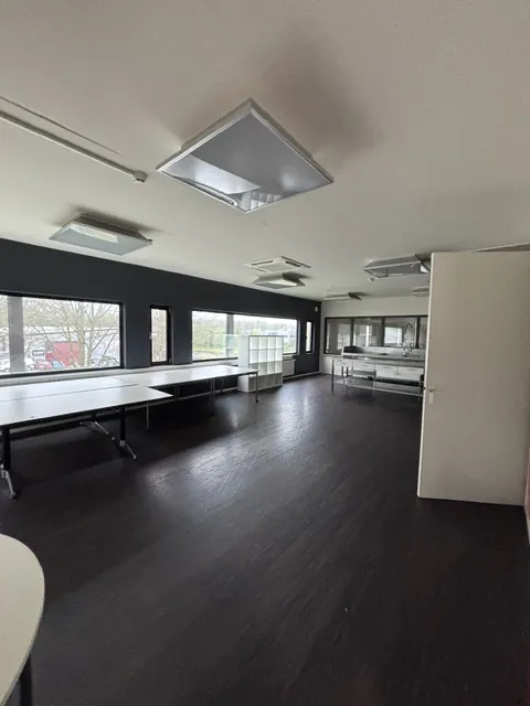 De lege kantine vóór de verbouwing, met een systeemplafond, een donkere vloer en losse tafels langs het raam