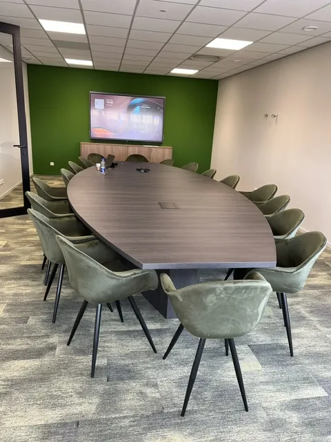 Vergaderruimte met een lange ovale tafel, olijfgroene stoelen en een scherm op een groene wand