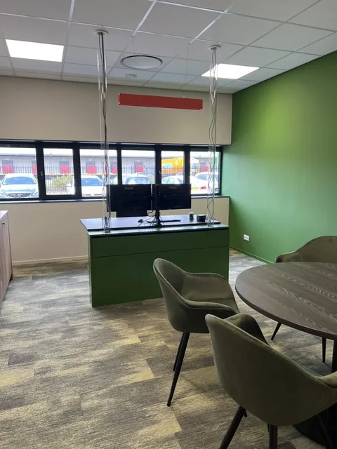 Werkplek met een groen bureaublok en twee schermen, naast een ronde overlegtafel met olijfgroene stoelen