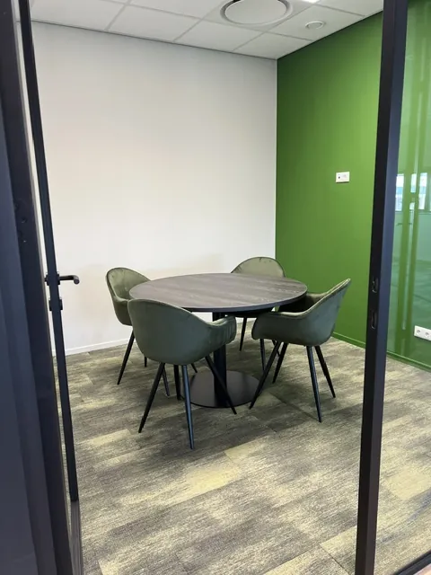 Overlegruimte achter glas met een ronde tafel, olijfgroene stoelen en een groene wand