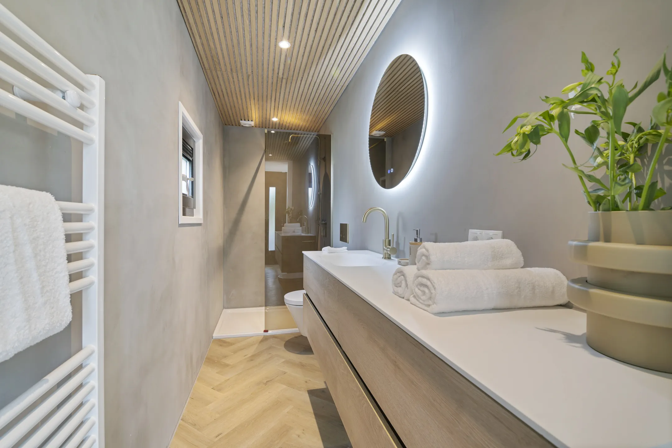 De badkamer met een lange wastafel, een ronde spiegel en een houten latjesplafond