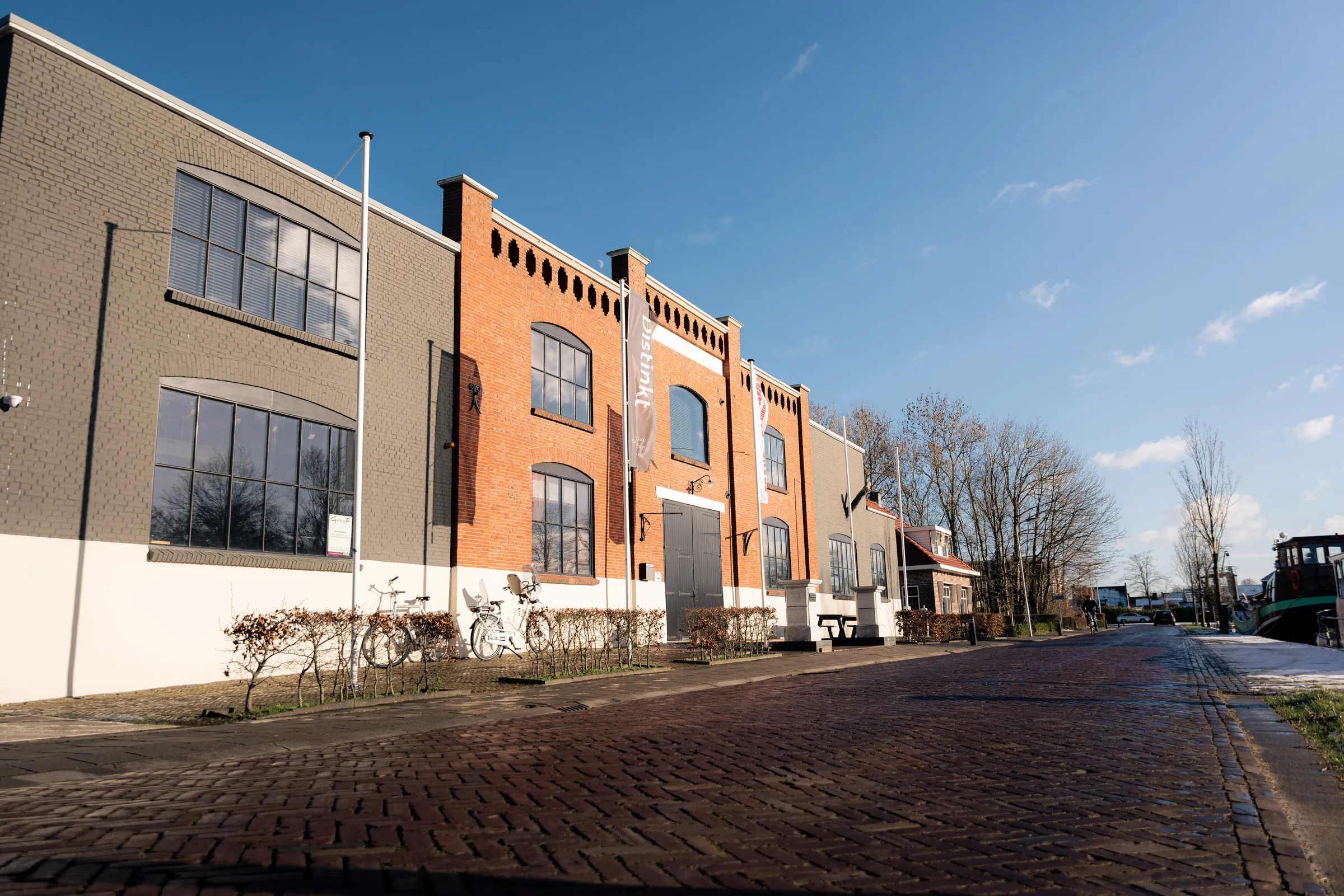 De rode bakstenen fabrieksgevel van FJ08 aan een klinkerstraat in Joure, onder een blauwe lucht