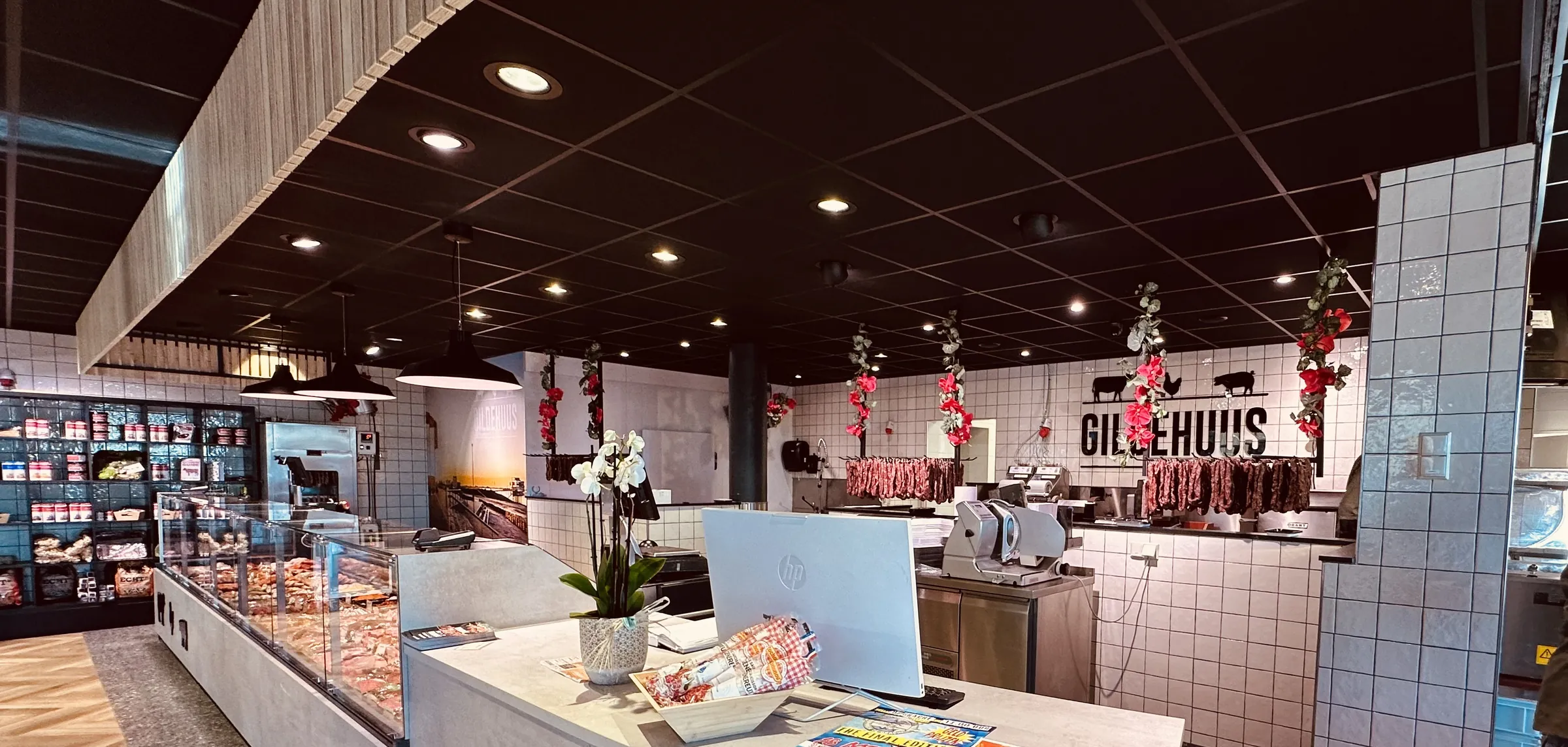 Balie met een orchidee en kassa voor een witte tegelwand met het Gildehuus-logo en hangende worsten