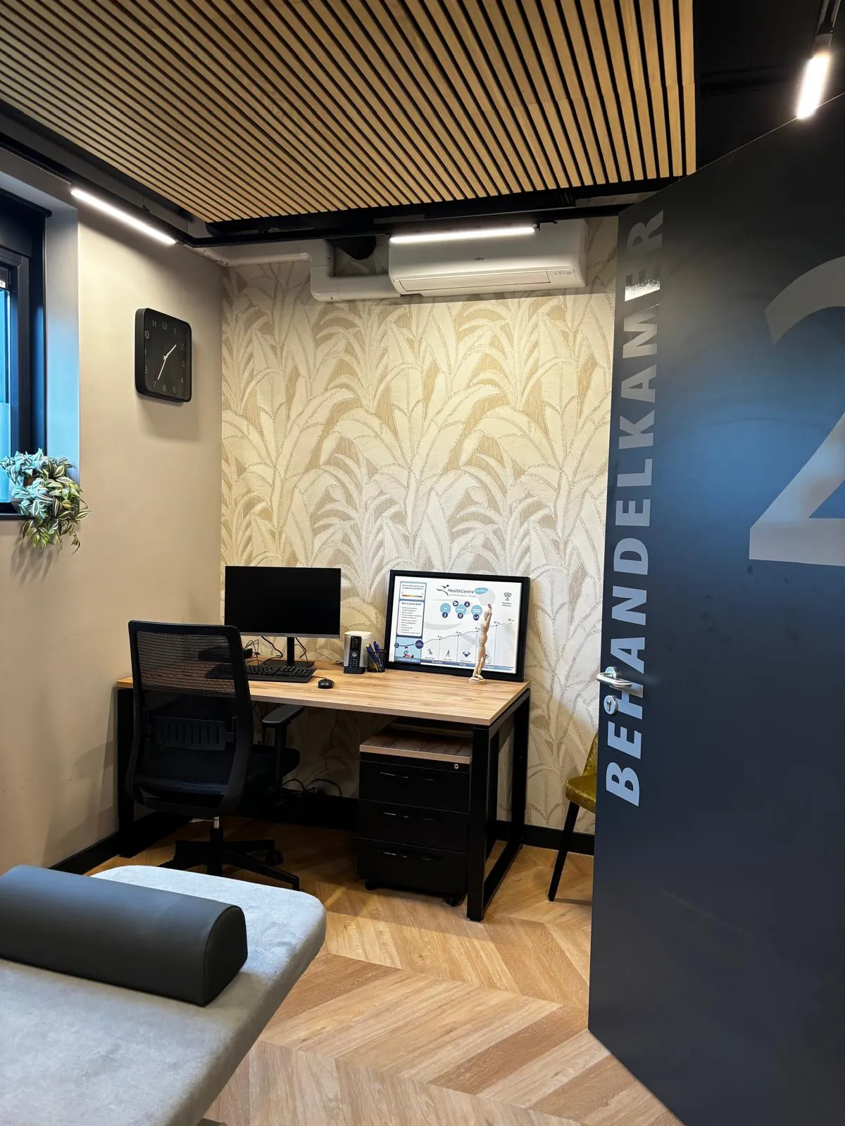 Behandelkamer met palmbladbehang, een bureau en een houten lamellenplafond, gezien vanuit de deur