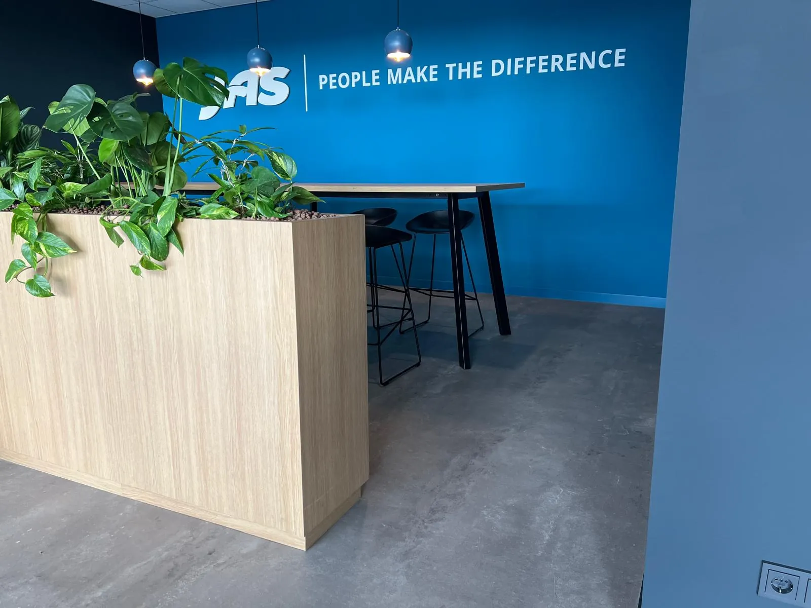 Pauzeplek met een statafel en barkrukken voor een felblauwe wand met het JAS-logo, naast een eiken plantenbak