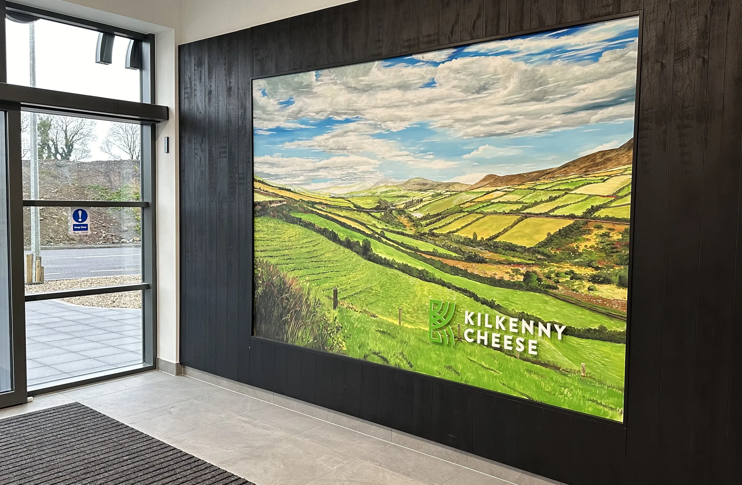 Entreehal met een ingelijst geschilderd heuvellandschap en het 3D-logo van Kilkenny Cheese op een zwarte houten wand