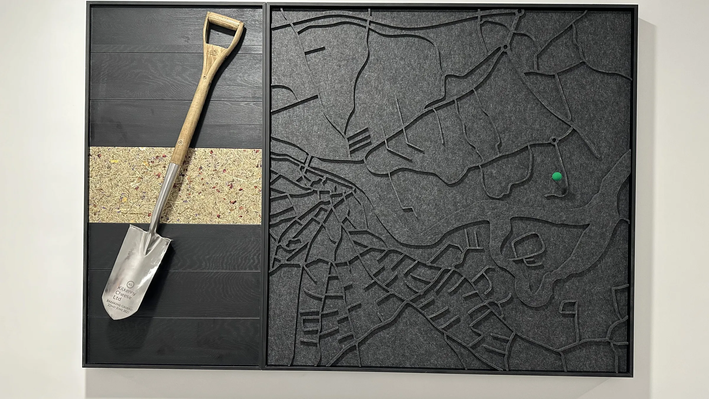 Wandkunstwerk met een gegraveerde schop op zwart hout naast een gelaserde vilten plattegrond met een groene pin
