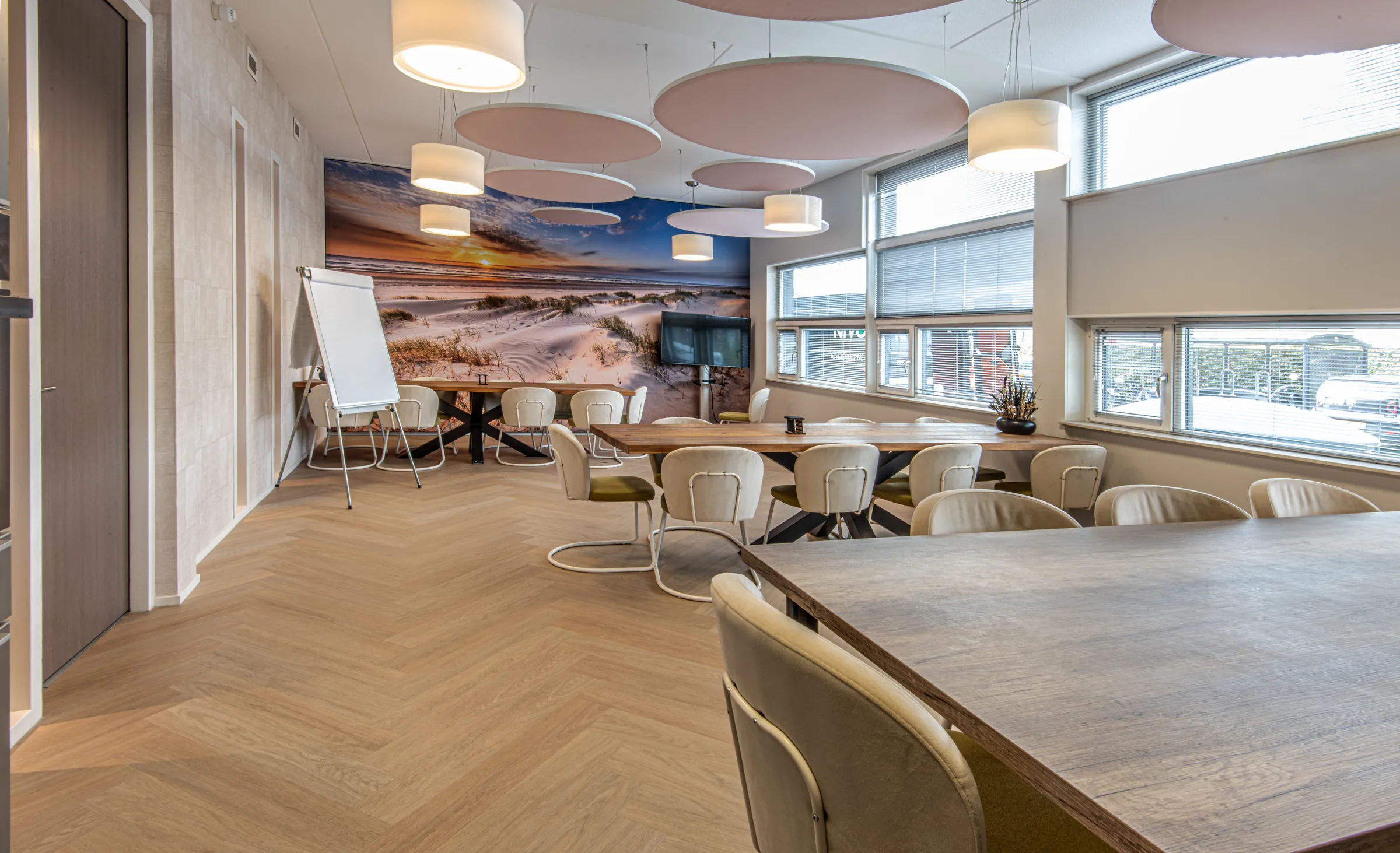 Overlegruimte met houten tafels, crèmekleurige stoelen, roze akoestische plafondeilanden en fotobehang van duinen bij zonsondergang