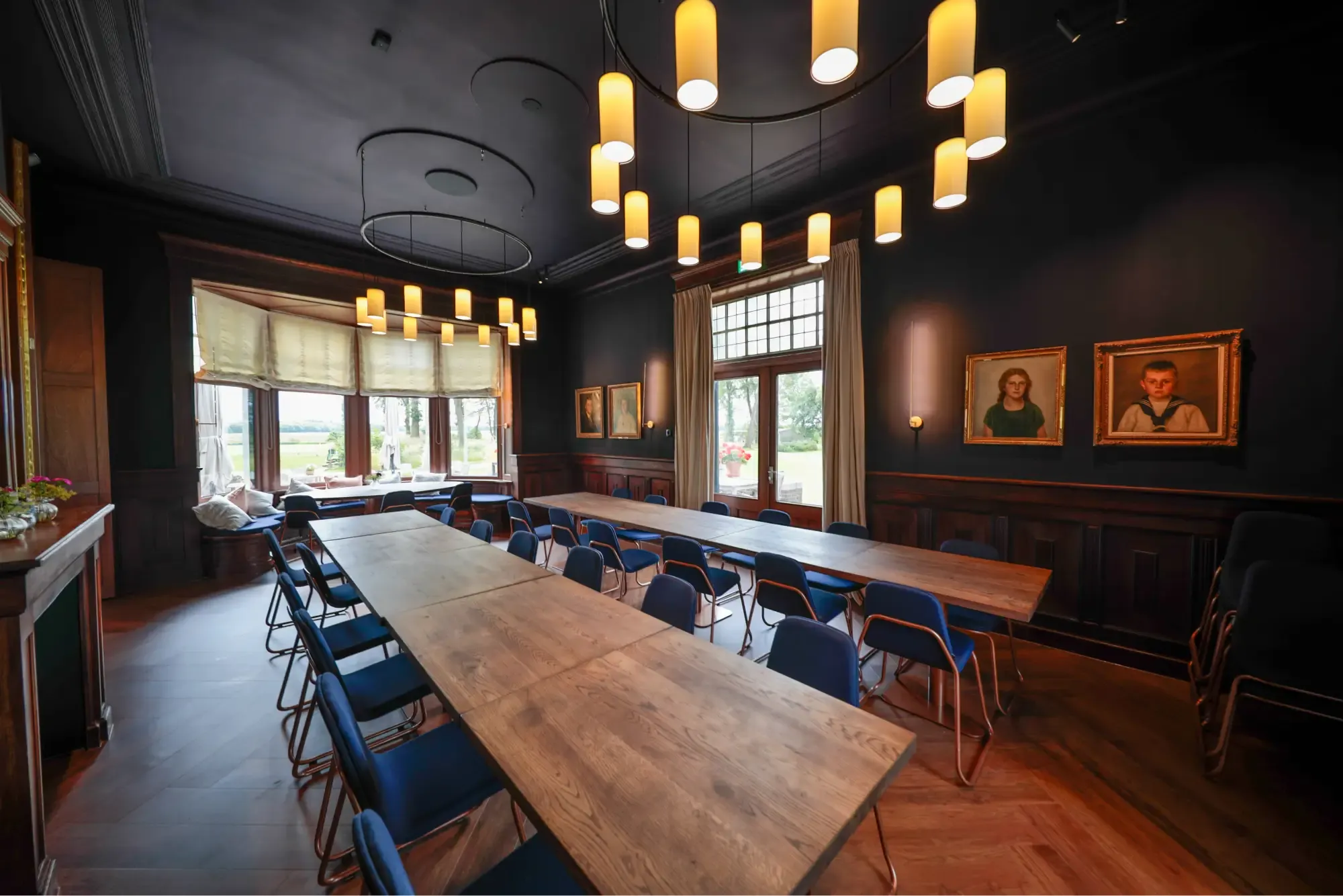 Een donkere zaal met houten lambrisering, een lange tafel en een ronde hanglamp