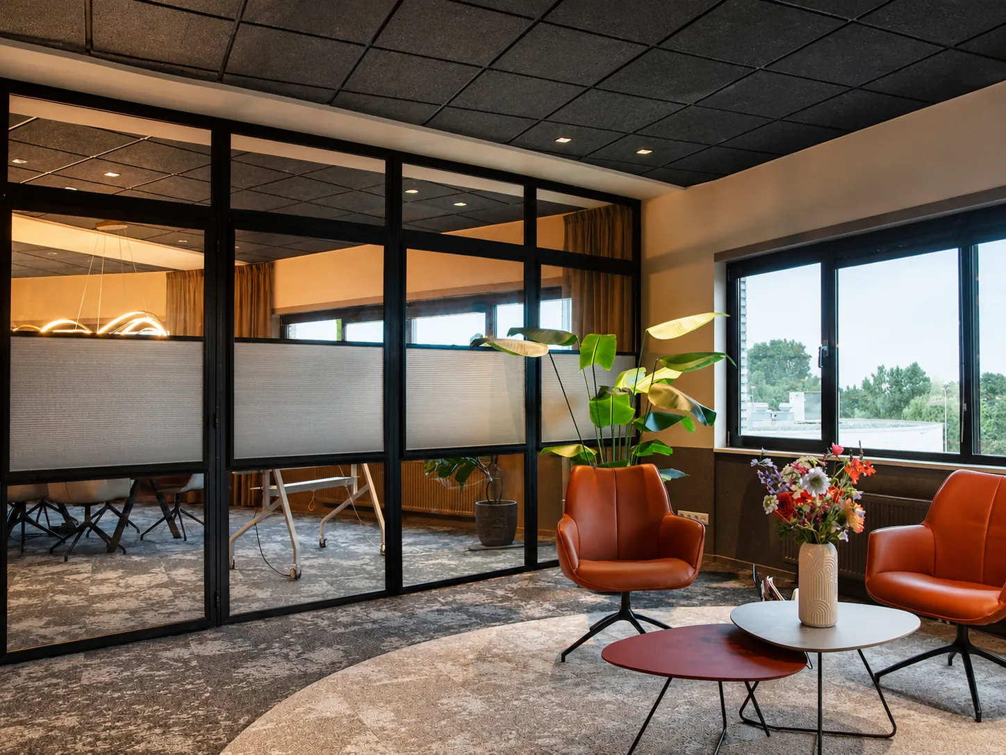 Zithoek met oranje fauteuils naast een zwart stalen glaswand met plissé, met zicht op de vergaderzaal