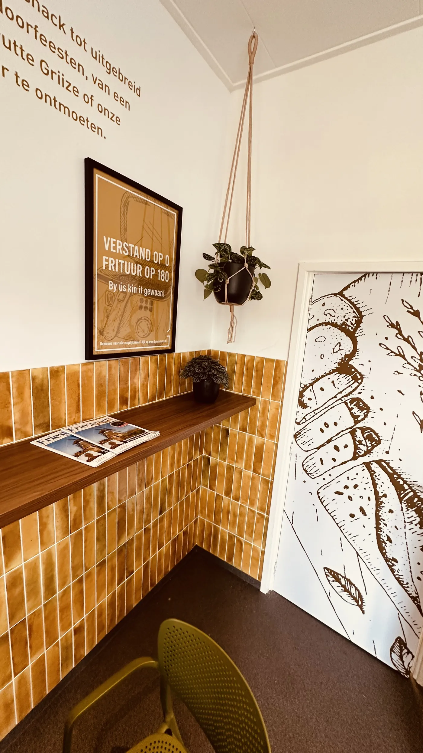 Een smalle houten wandplank tegen de gele tegels, met een poster erboven