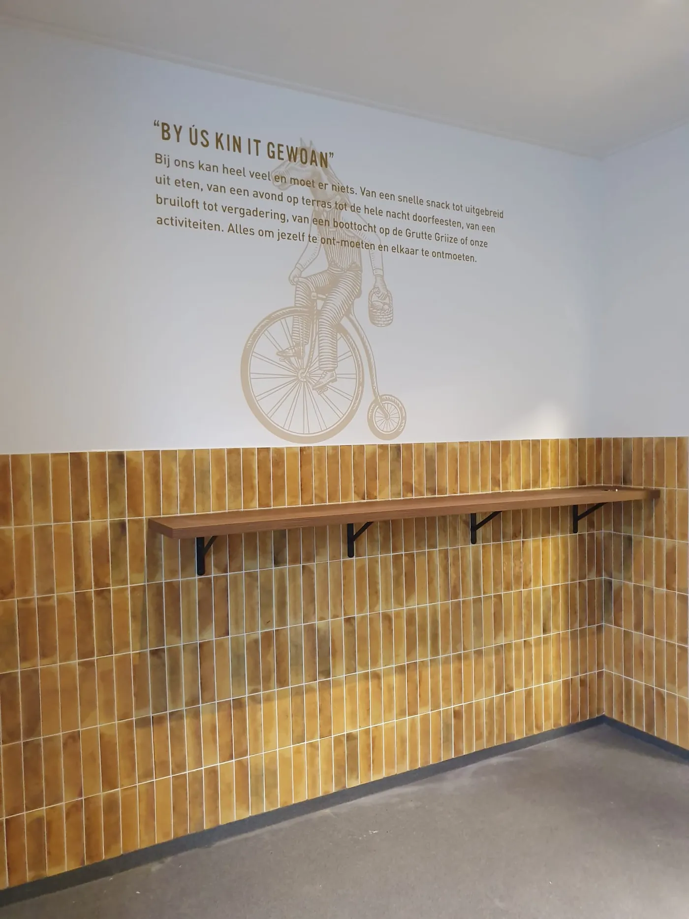 Een tekening van een paard op een hoge fiets op de muur van Snackbar De 2 Gemeenten, met de tekst By ús kin it gewoan