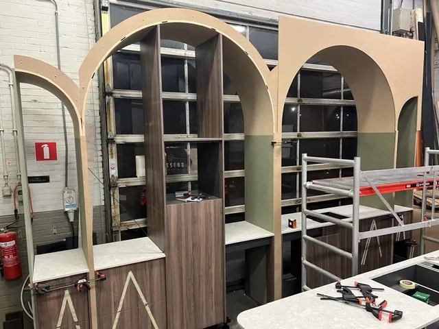 Boogvormige vitrinekasten in aanbouw in de werkplaats, met lijmklemmen en een rolsteiger
