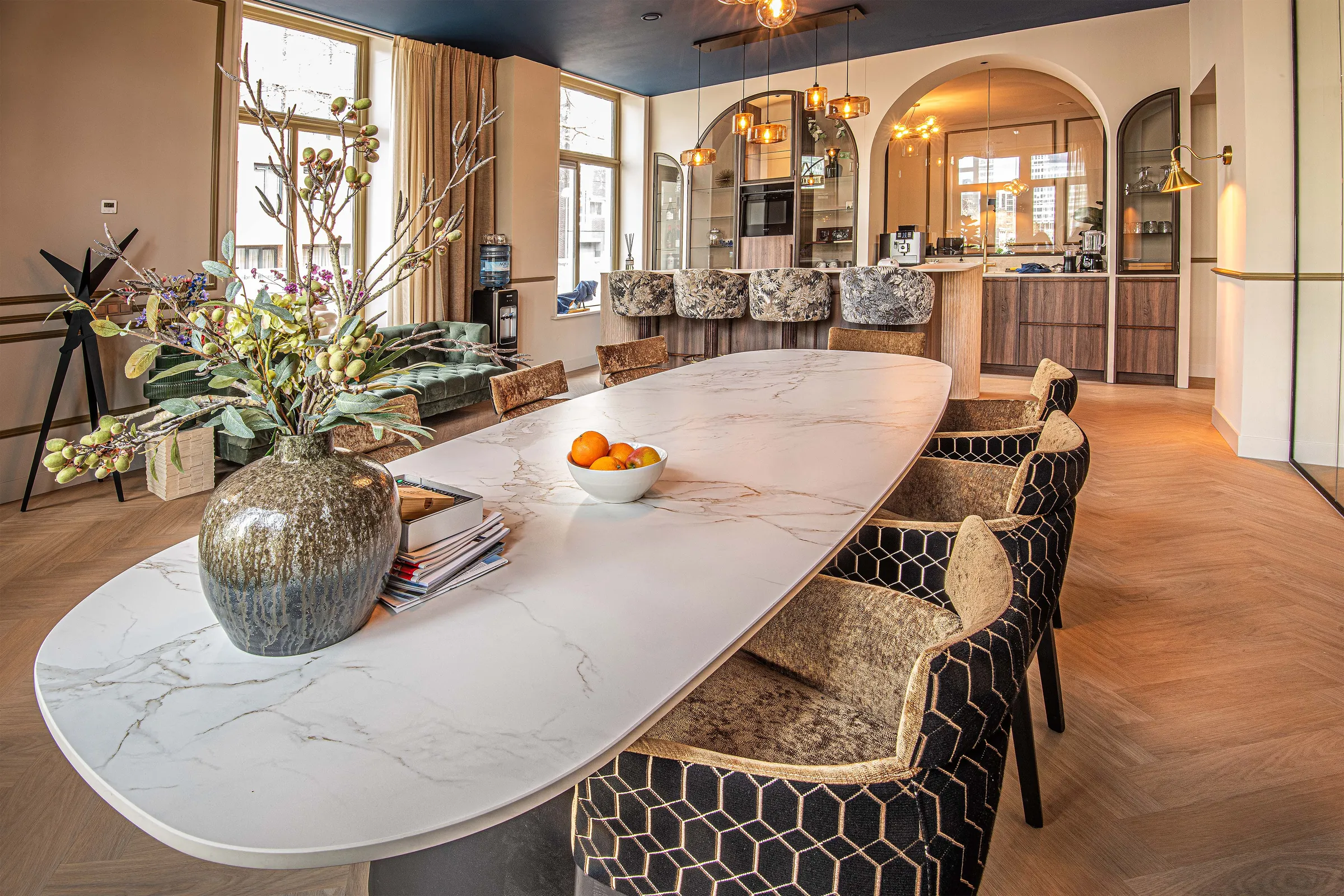 Lange ovale tafel met marmerlook blad en gestoffeerde stoelen, met daarachter de bar en de boogkasten