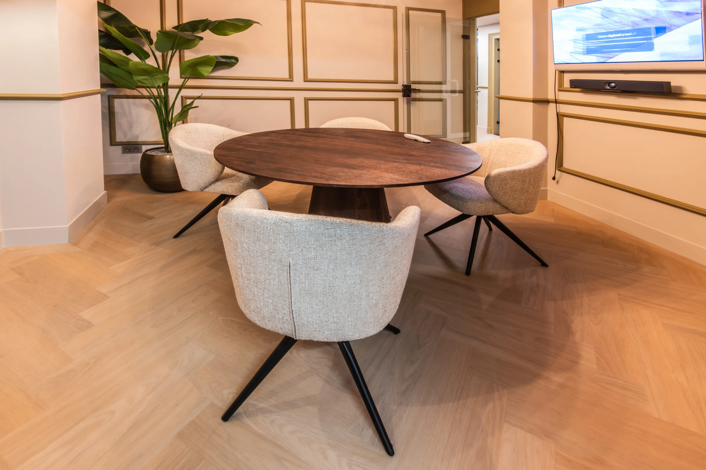 Overleghoek met een ronde donkere houten tafel, crèmekleurige stoelen en wandlijsten met goudkleurige profielen