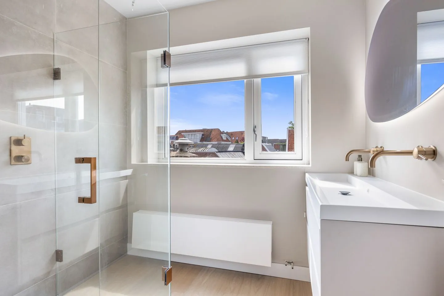Badkamer met een glazen inloopdouche, koperkleurige kranen, een witte wastafel en een organisch gevormde spiegel met ledrand