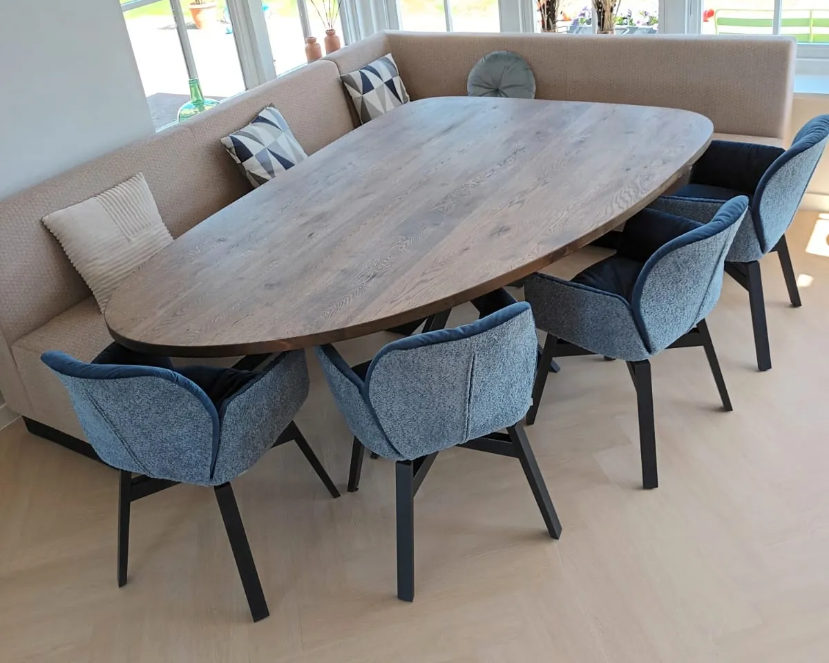 Een ovale houten eettafel met blauwe stoelen in de erker, met ramen aan drie kanten